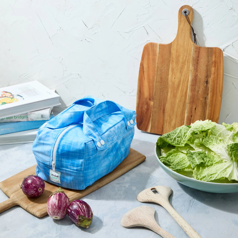 Blue reusable produce bag on a kitchen counter with a bowl of lettuce, cutting board, and baby eggplants.