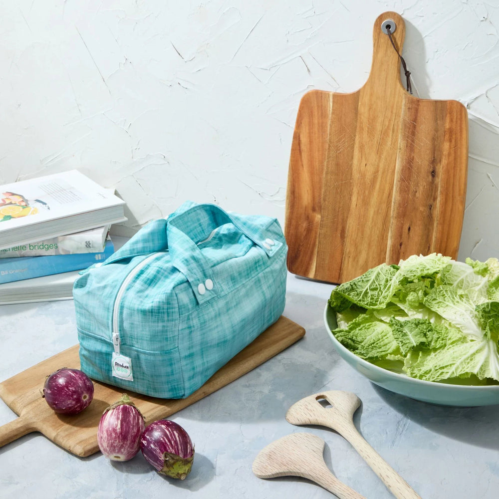 Teal reusable produce bag on a kitchen counter with a bowl of lettuce, cutting board, and baby eggplants.