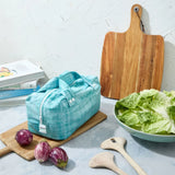 Teal reusable produce bag on a kitchen counter with a bowl of lettuce, cutting board, and baby eggplants.