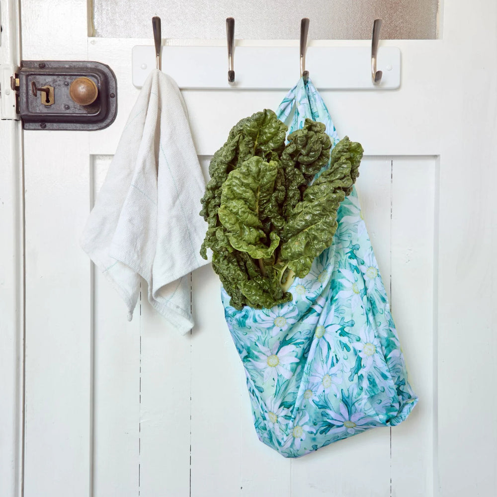Green leafy vegetables in a reusable shopper bag hanging on a white door with hooks.