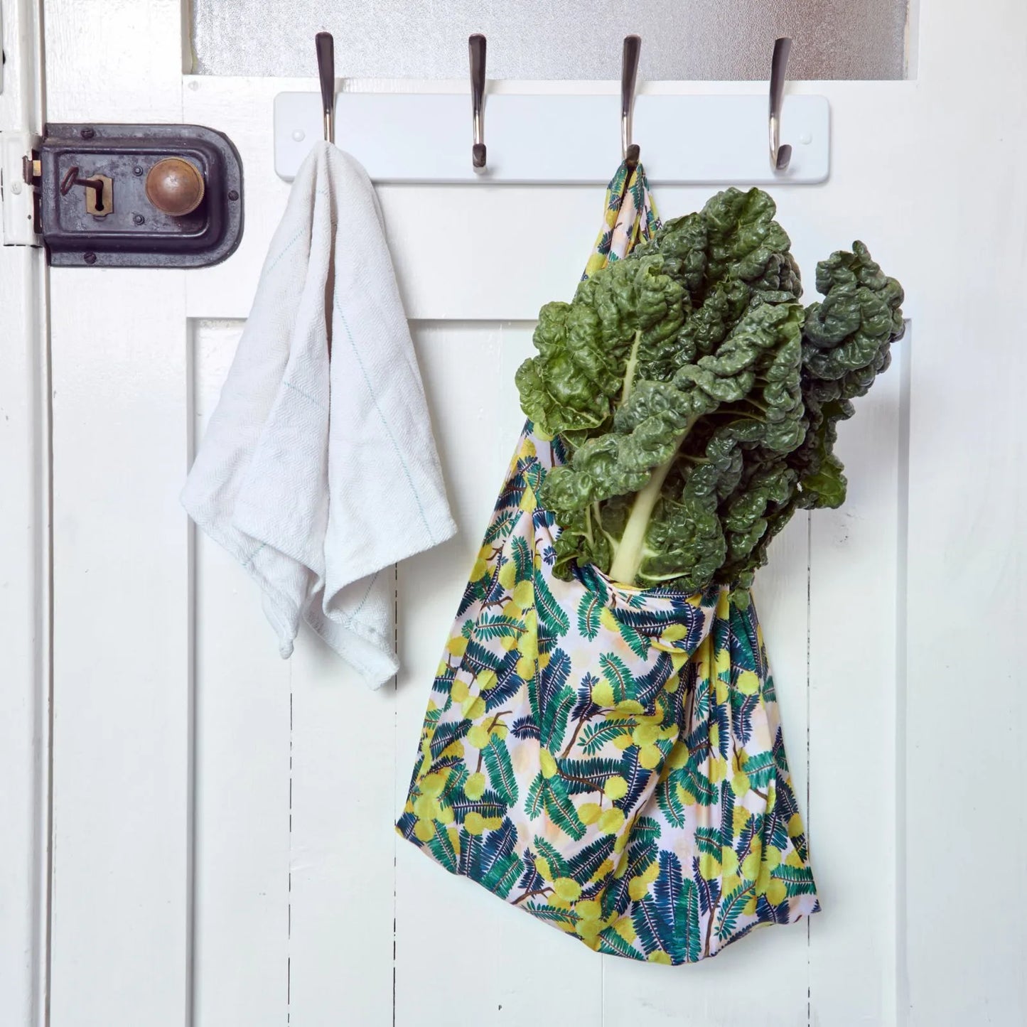 Green leafy vegetables in a reusable shopper bag hanging on a white door with hooks.