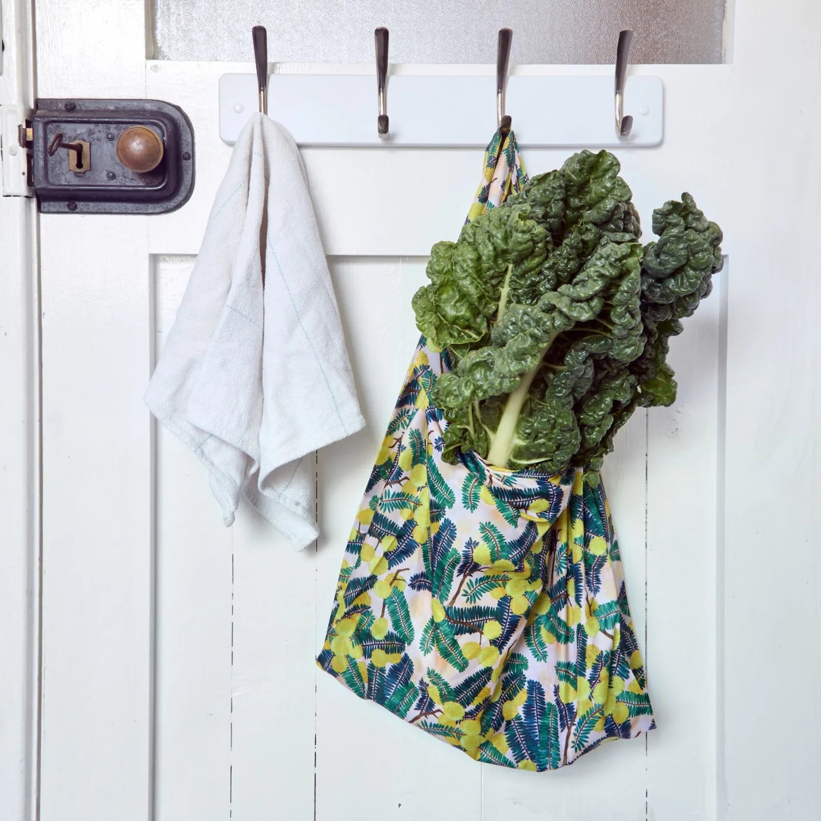 Green leafy vegetables in a reusable shopper bag hanging on a white door with hooks.