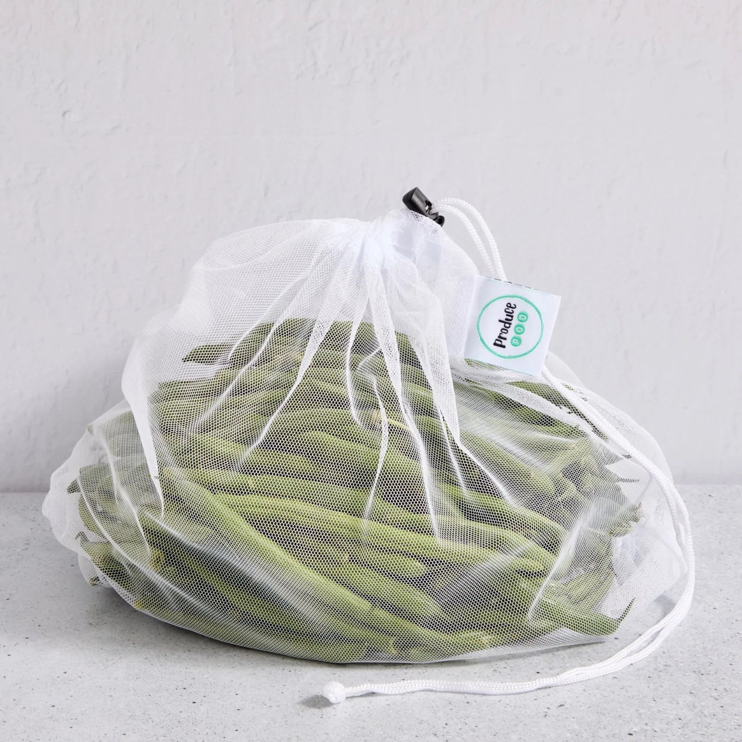 White drawstring mesh reusable produce bag filled with green beans on a light grey background